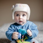 cute baby boy 6 months old looking at camera. He is wearing a helmet to correct plagiocephaly and he is playing with a green toy while wearing a blue jaket